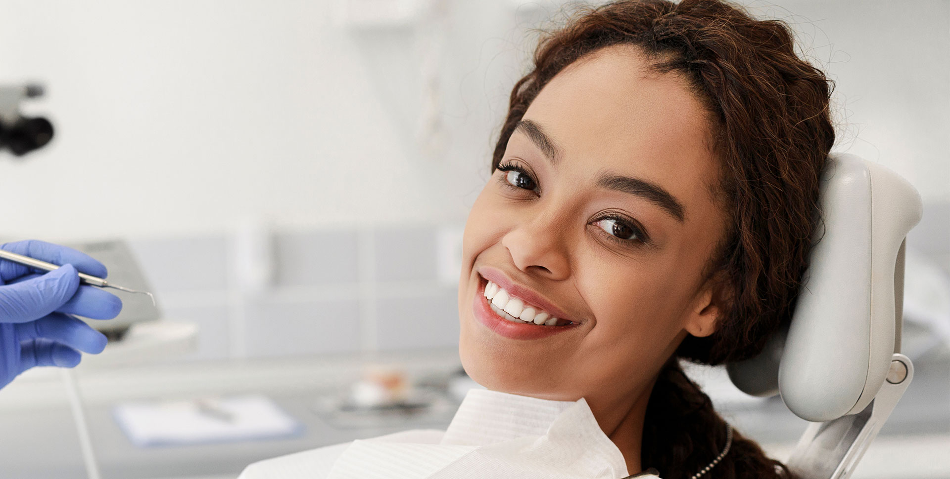 A woman is smiling while sitting in a dental chair with her eyes closed, and a dental professional appears to be performing a procedure on her teeth.