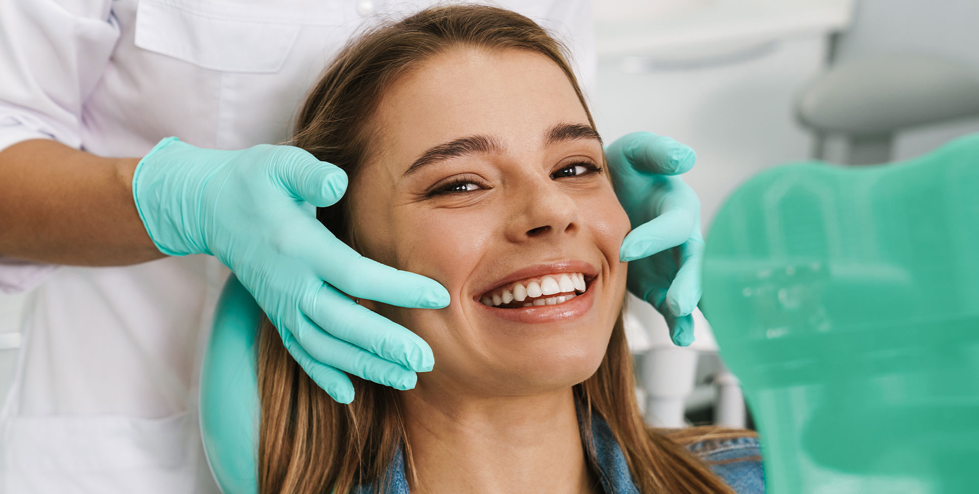 The image shows a young woman smiling while sitting in a dental chair, receiving dental care from a professional wearing gloves and a face mask.