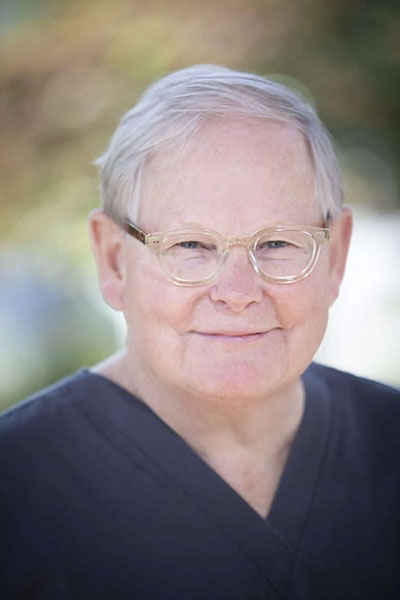 The image shows an older man wearing glasses, with white hair and a beard, dressed in a black shirt. He is smiling and appears to be outdoors during the daytime.