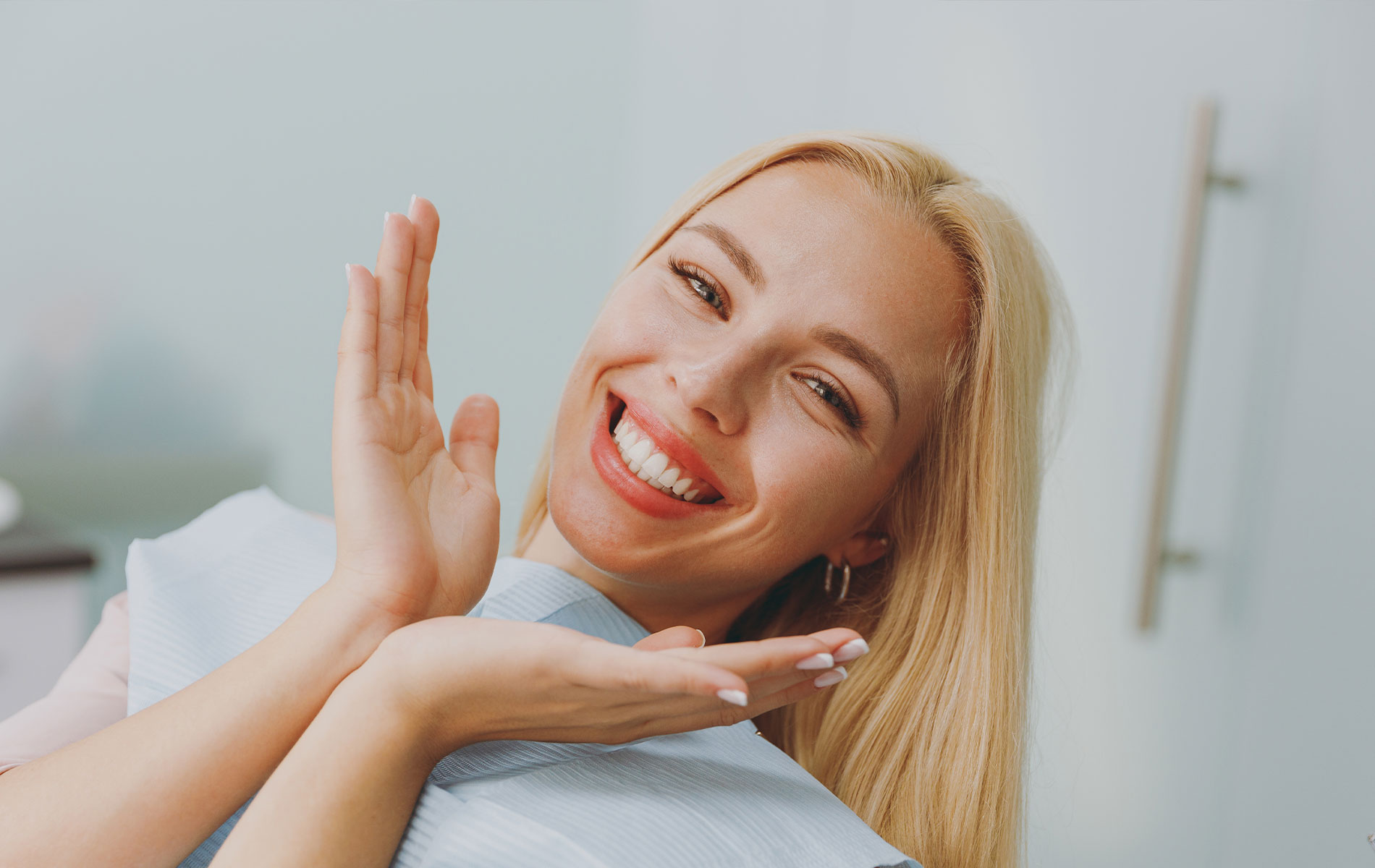 The image shows a woman with blonde hair, smiling broadly, with her hands raised to her cheeks in a gesture of excitement or surprise. She is wearing a white dental hygiene mask and appears to be seated in a dental office setting.
