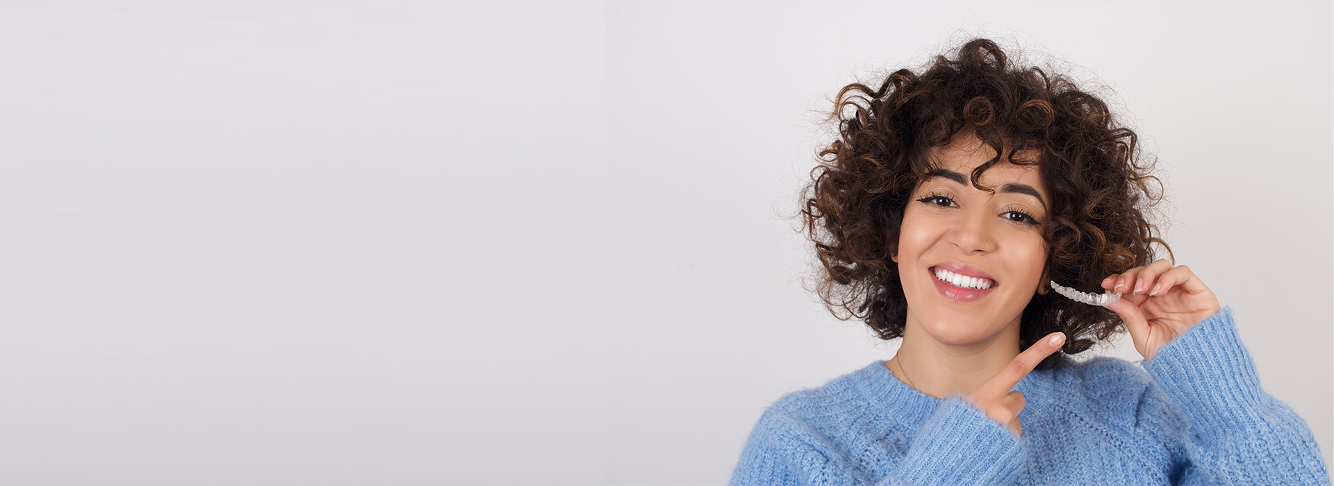 A smiling woman with curly hair, holding a smartphone to her ear, against a plain background.