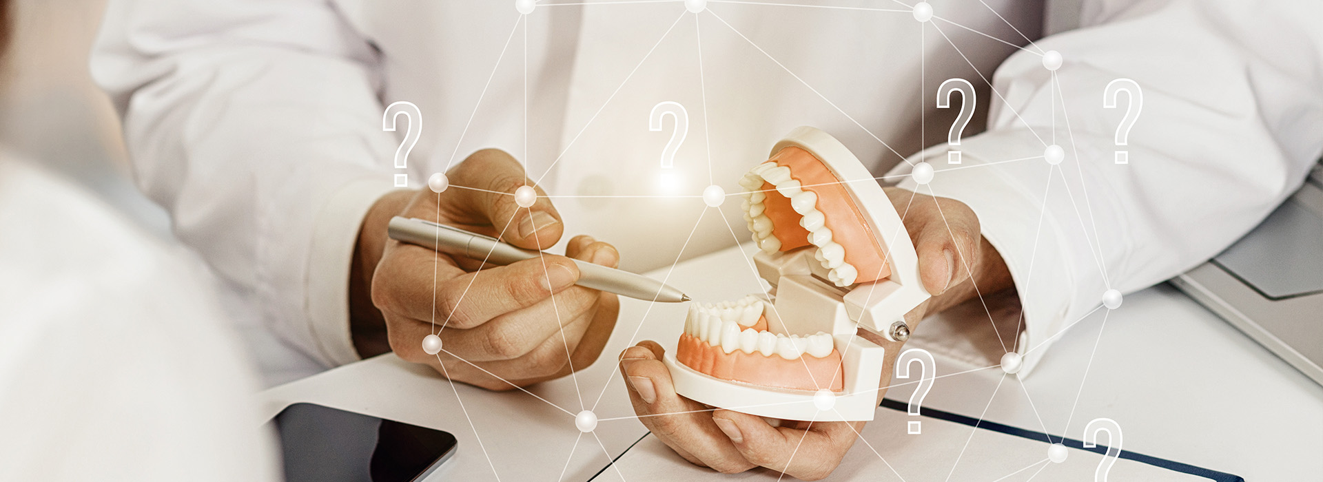 The image is a composite photograph featuring an individual s hands holding a model of a human mouth with teeth, set against a background that includes a digital overlay displaying various icons and numbers, as well as a partial view of another person wearing a white lab coat.