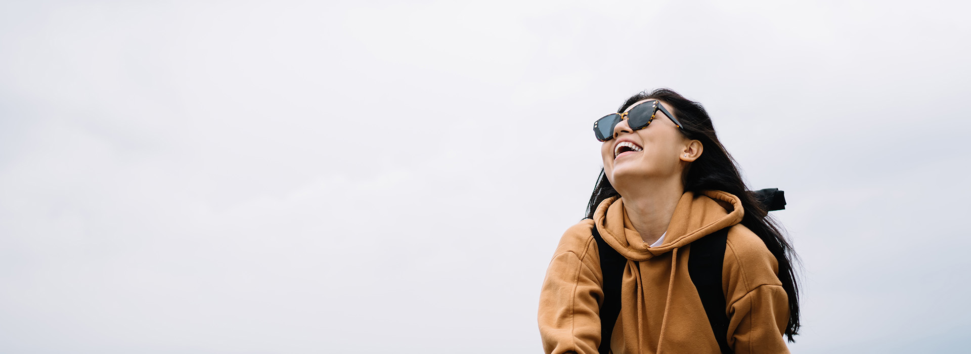 A person, possibly a woman, is standing outdoors in a light-colored hoodie and sunglasses, smiling and looking away from the camera.