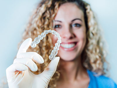 A woman in a blue shirt is holding up a large, transparent dental retainer with her right hand.