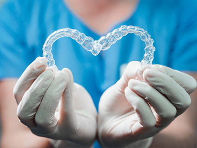 An image of a person s hands holding a heart-shaped object that appears to be a transparent dental appliance, with a background that suggests a medical setting.