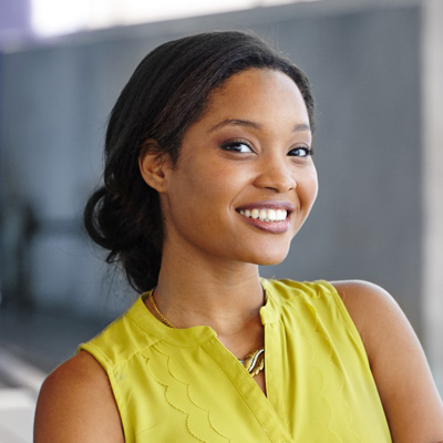 A young woman with a bright smile is posing for the camera, wearing a yellow blouse and holding her hair up. She stands against a backdrop of a building wall.