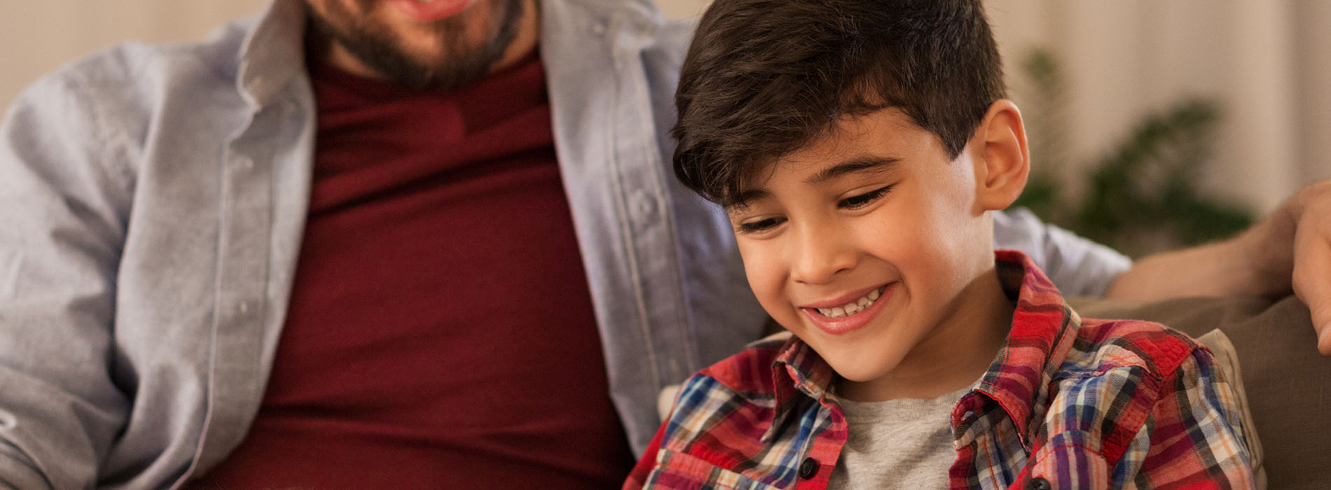 The image shows a man and a young boy, both smiling, sitting together on a couch with a laptop in front of them.