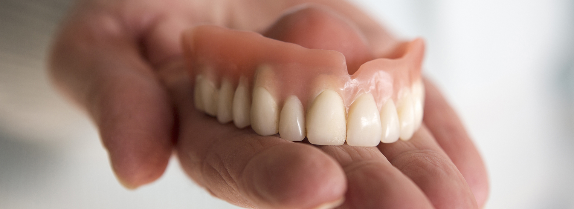 The image features a hand holding an upper denture with teeth, against a blurred background.