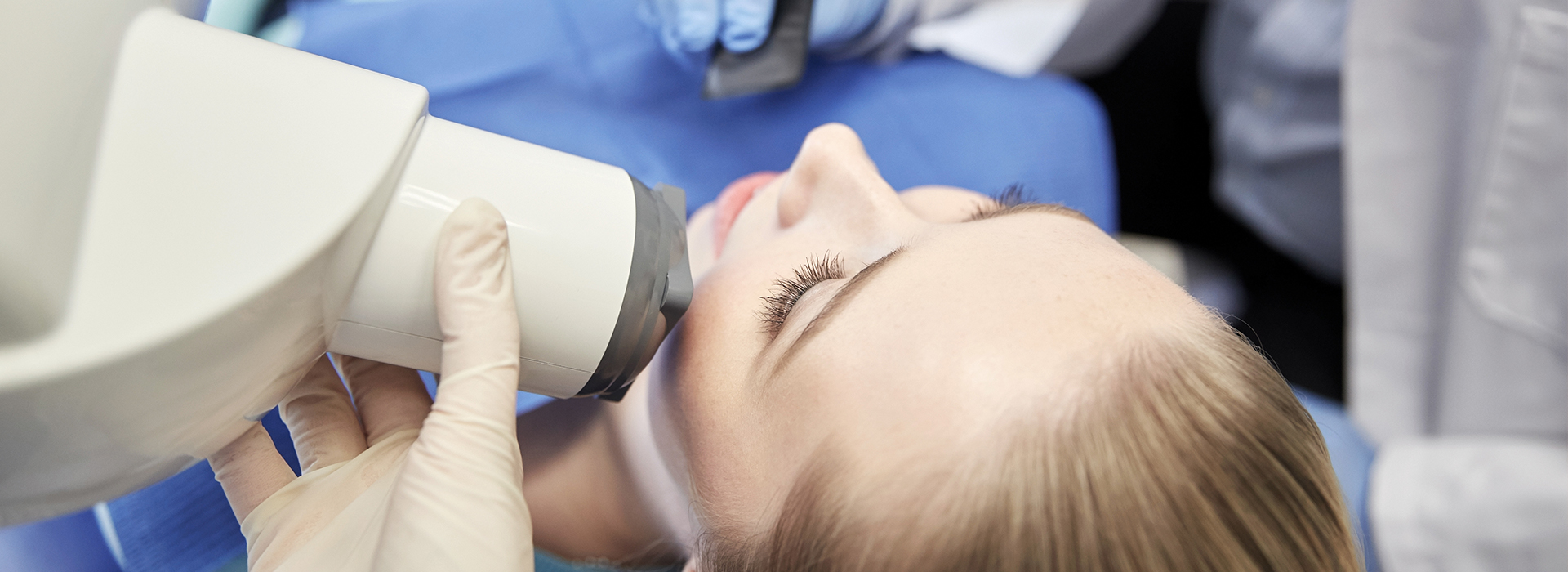 The image shows a person lying down with their head in a dental or medical device, likely a machine used for dental procedures or medical examinations, while another person is performing the procedure.