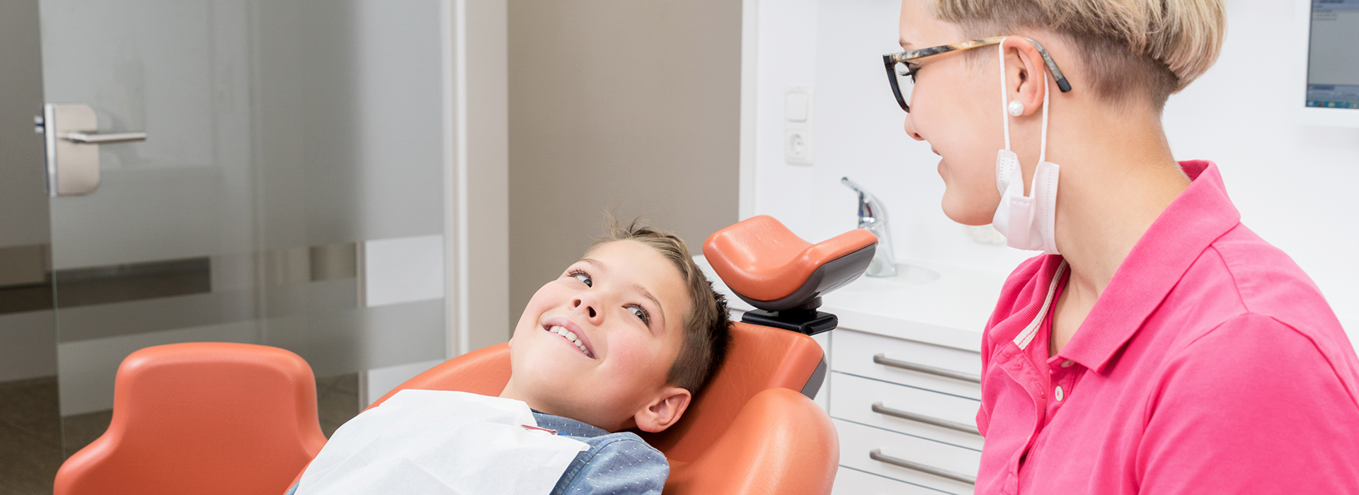 The image shows a dental office setting where a child is sitting in the dentist s chair, receiving care, with a female dental professional standing beside him.