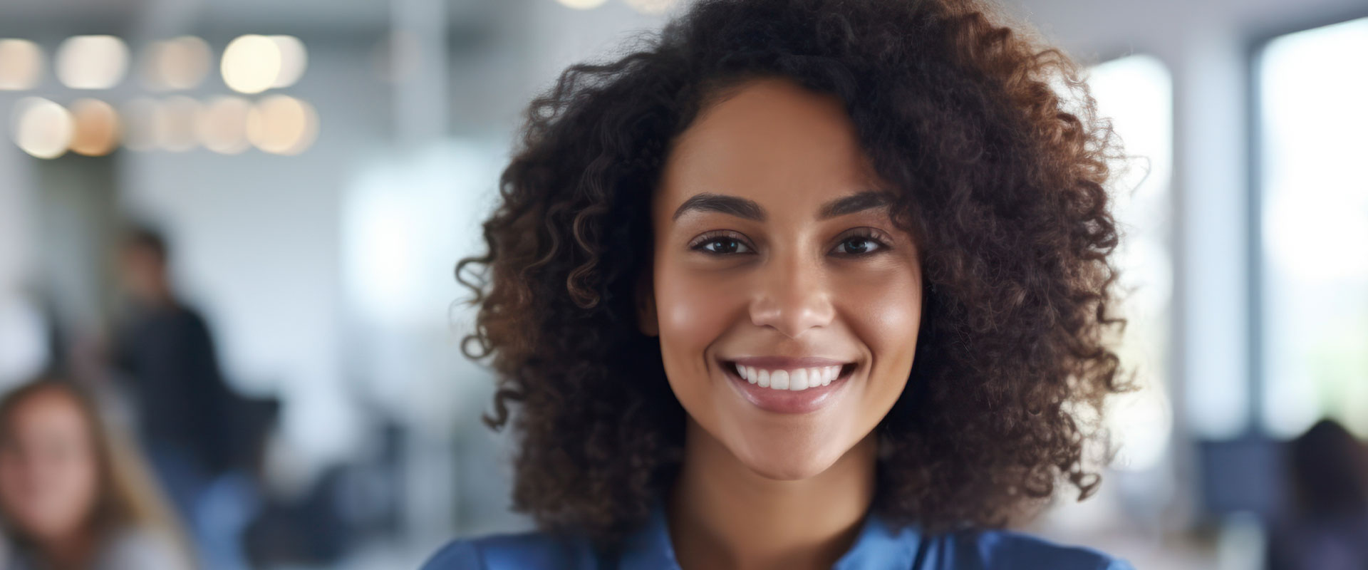 The image shows a woman with curly hair, smiling at the camera. She appears to be in an office setting, possibly presenting or engaging with others.