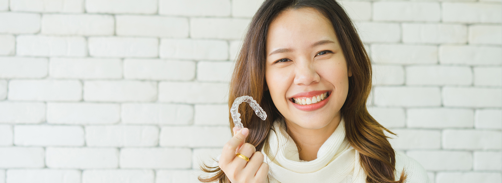 A woman with a smile, holding a ring, against a brick wall background.