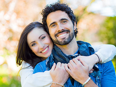 A man and a woman are hugging each other in an outdoor setting, both smiling and looking content.