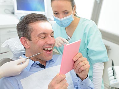 A man is seated in a dental chair, holding up a pink card with a surprised expression while being attended to by a dentist who is smiling and looking at the card.