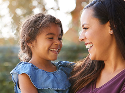 A young child and an adult woman smiling at the camera, with a blurred background that suggests they are outdoors during daylight.