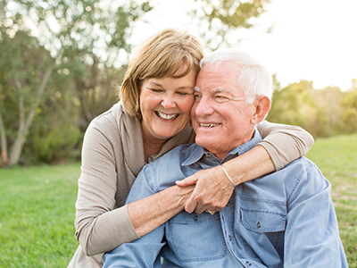 An elderly couple embracing outdoors, with the man wearing a blue shirt and the woman in a white top.