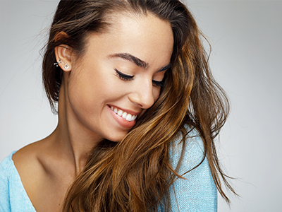 A smiling woman with long hair, wearing a light-colored top and earrings, against a neutral background.