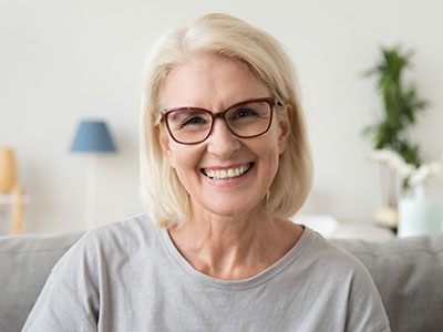 The image shows a woman with short blonde hair, wearing glasses and a light-colored top, smiling at the camera.