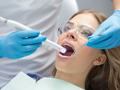 The image shows a person receiving dental treatment, with a dental professional using a device to examine their teeth while the patient is seated in a dentist s chair.