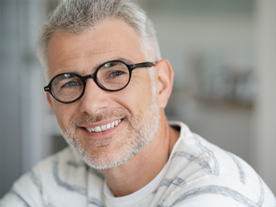 The image is a portrait of a smiling man with short gray hair, wearing glasses and a white shirt, against a blurred background.