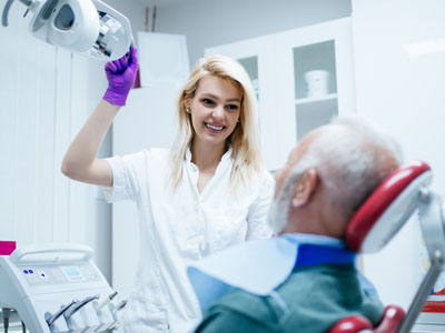 A woman in a white coat, holding a dental mirror with a smile on her face, stands next to an elderly man in a dentist s chair, who is wearing a surgical mask and receiving dental care.