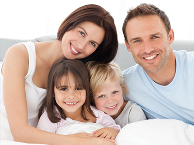 A family of four, including a man, woman, and two young children, posing together on a bed with smiles.