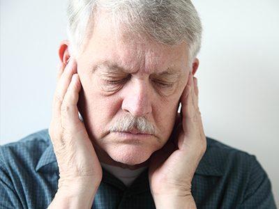 The image shows an elderly man with a concerned expression, holding his face in his hands.